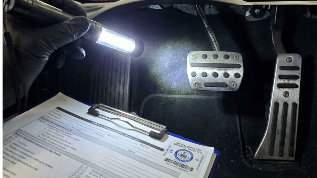 自動車の車検場で検査員が懐中電灯で運転席の足元を照らし、ペダルカバーの固定強度や滑り止め加工を厳しくチェックしている視点。