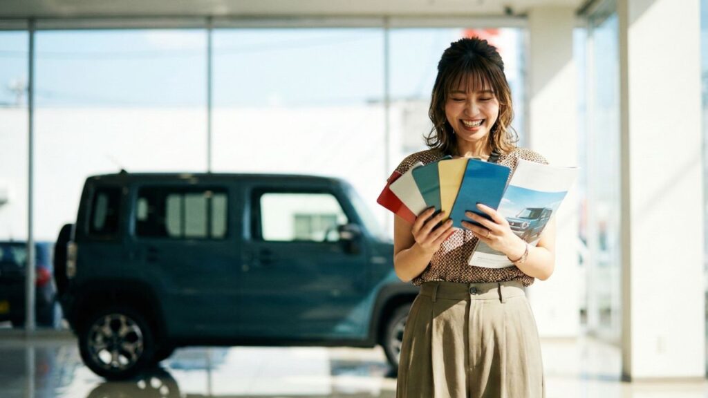 : カーディーラーのショールームで、カラフルなボディカラー見本やカタログを笑顔で見比べている日本人女性。